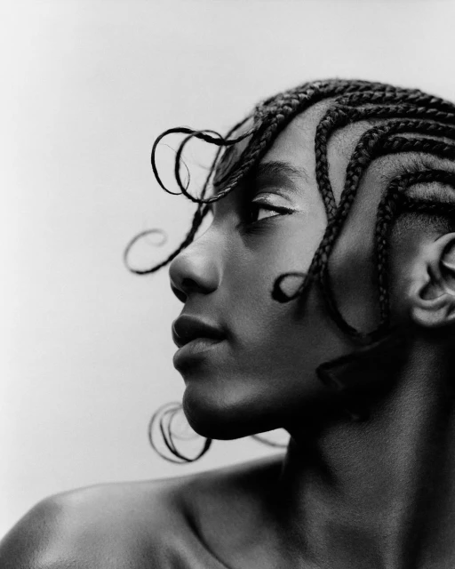 Striking black and white portrait of a woman with cornrow braids looking left, enhanced by bright eye makeup and against a neutral background.
