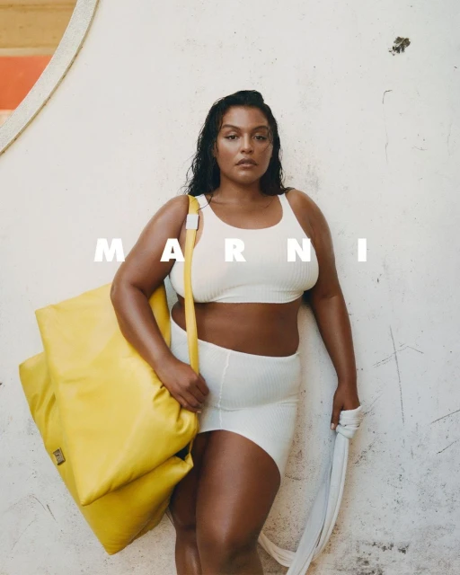 Woman poses with a yellow tote bag and a white towel. She wears a white crop top and shorts set against a textured white wall.