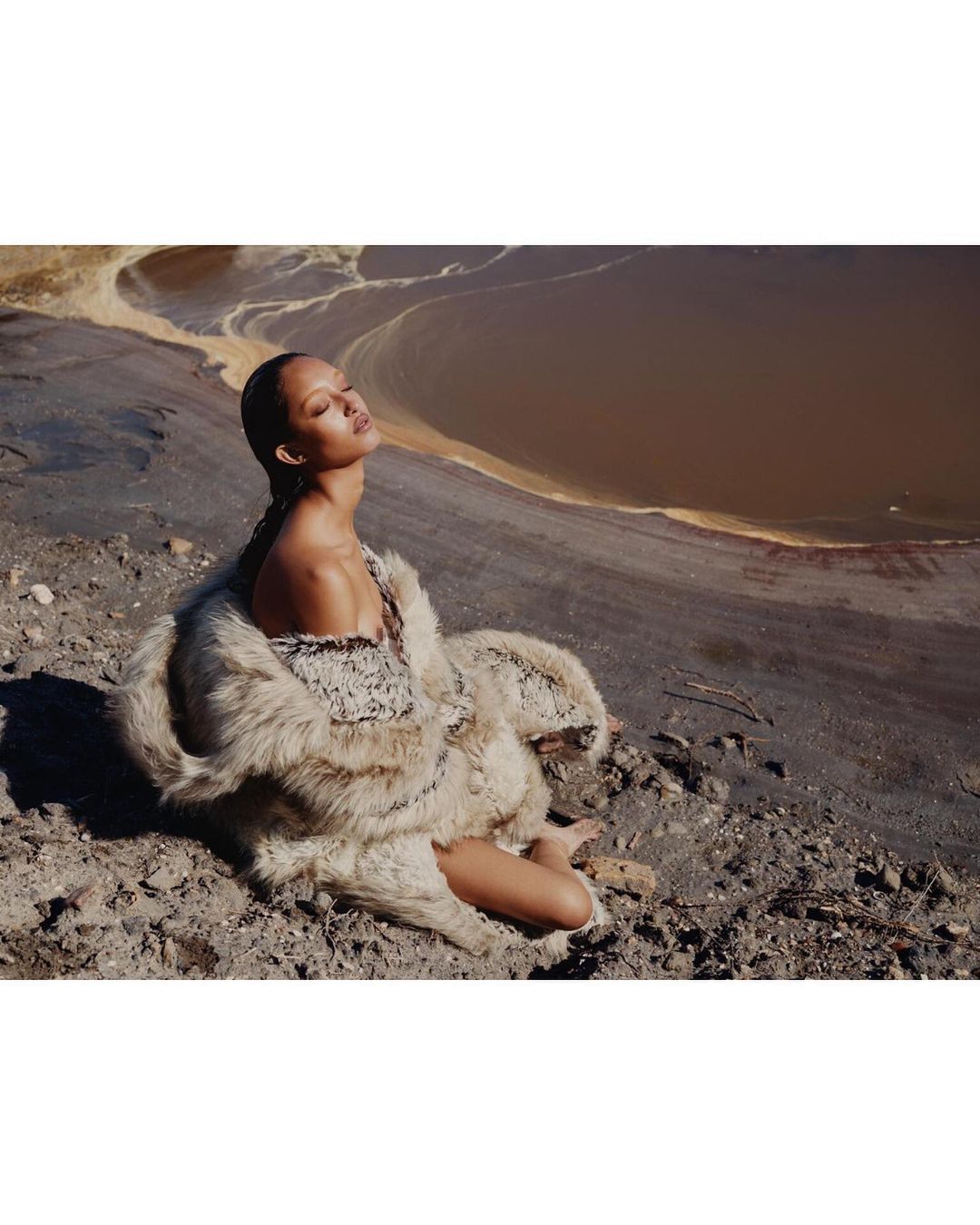 Woman in fluffy textured coat seated outdoors on rough terrain near muddy water by Tyler Mitchell