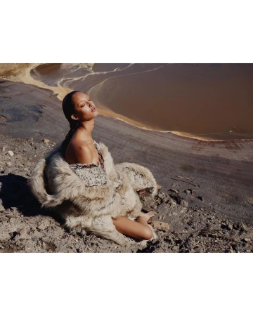 Woman in fluffy textured coat seated outdoors on rough terrain near muddy water by Tyler Mitchell