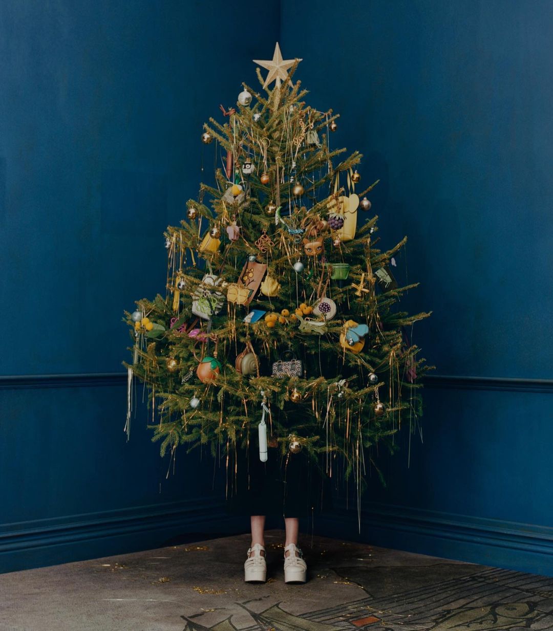 Decorated Christmas tree with star topper, ornaments, and tinsel in a room with dark blue walls and a patterned floor. White platform shoes visible.