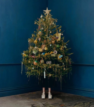 Decorated Christmas tree with star topper, ornaments, and tinsel in a room with dark blue walls and a patterned floor. White platform shoes visible.