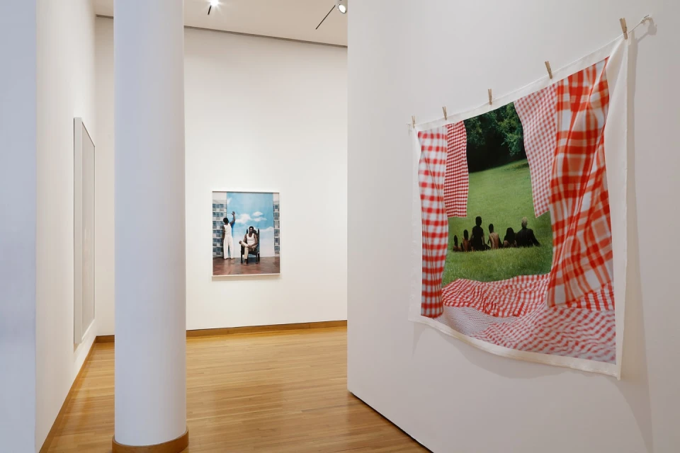 Installation view of photographic prints on display in an art gallery; a group of people sitting on grass in a park.