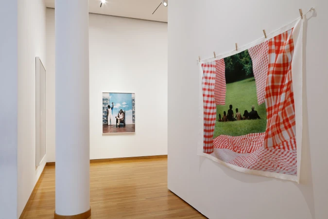 Installation view of photographic prints on display in an art gallery; a group of people sitting on grass in a park.