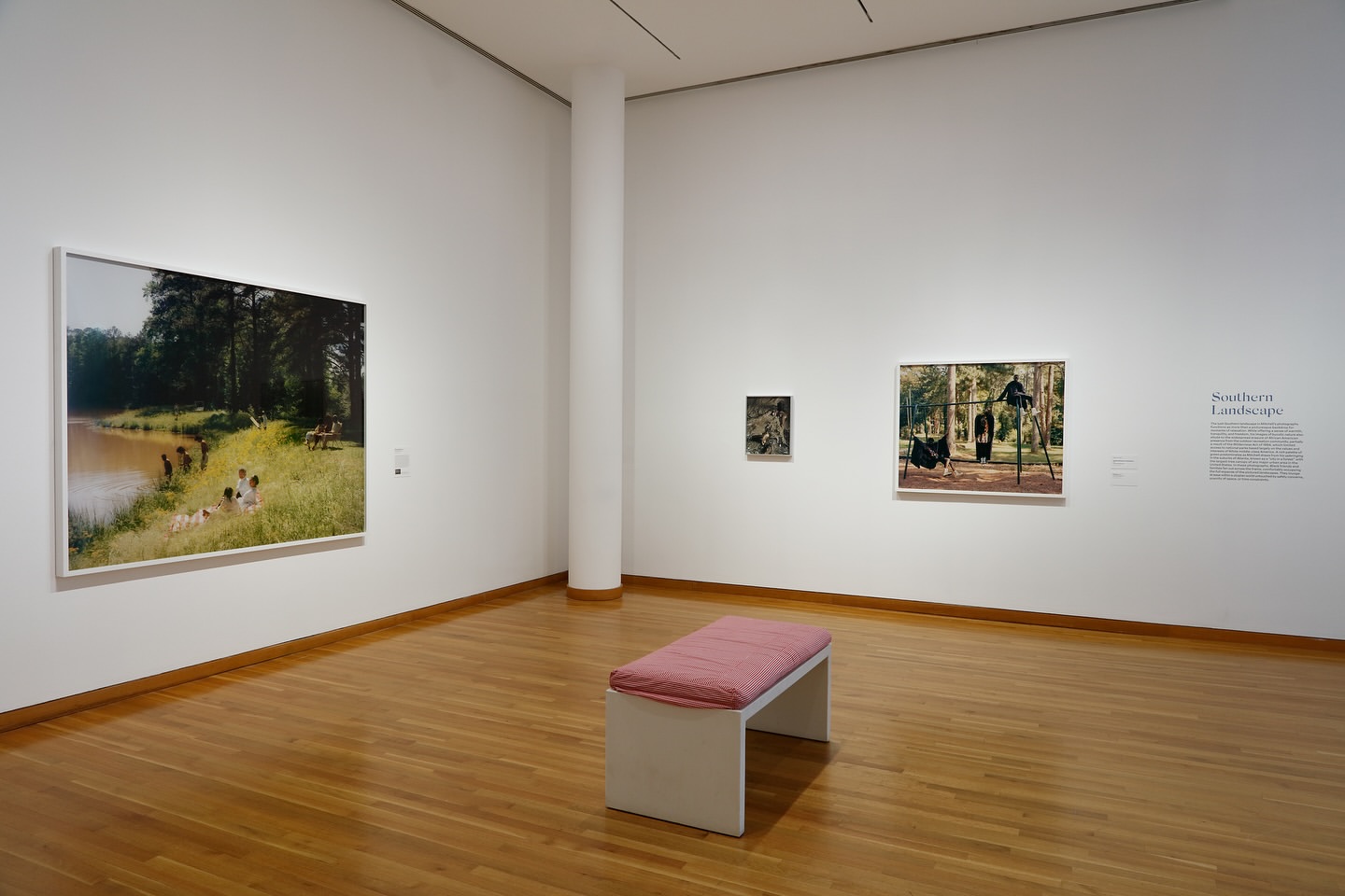 Art gallery view with paintings of outdoor scenes on the walls, a white bench, and a hardwood floor in a well-lit space.