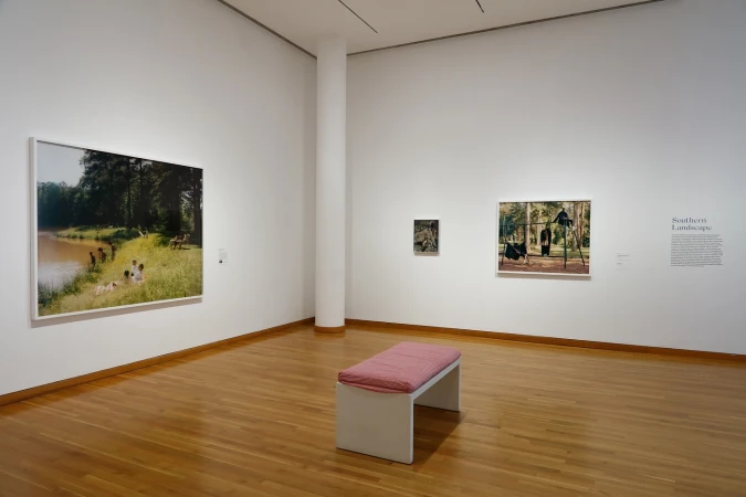Art gallery view with paintings of outdoor scenes on the walls, a white bench, and a hardwood floor in a well-lit space.