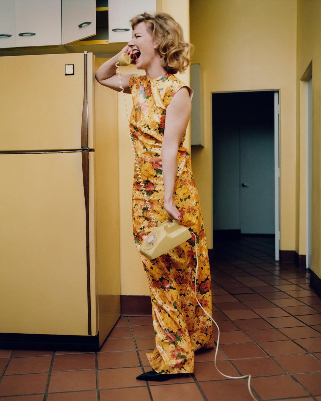Woman in floral dress talks on a vintage yellow landline phone in a yellow kitchen with brown tiled floor, with a yellow fridge.