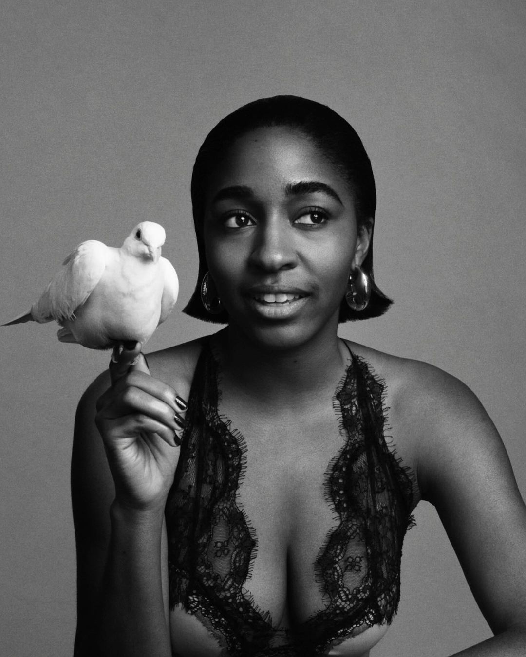 Black and white portrait of an African American woman holding a white bird, wearing a lace top, and statement earrings against a grey backdrop.