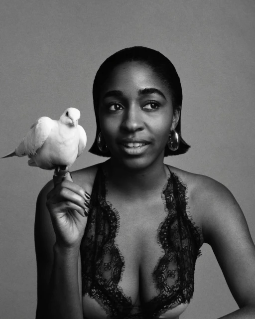 Black and white portrait of an African American woman holding a white bird, wearing a lace top, and statement earrings against a grey backdrop.