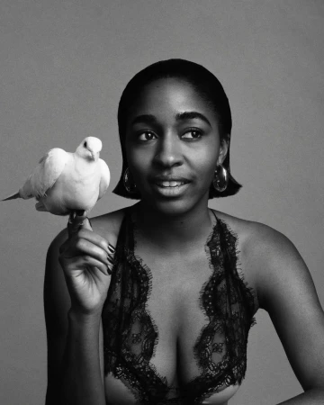 Black and white portrait of an African American woman holding a white bird, wearing a lace top, and statement earrings against a grey backdrop.