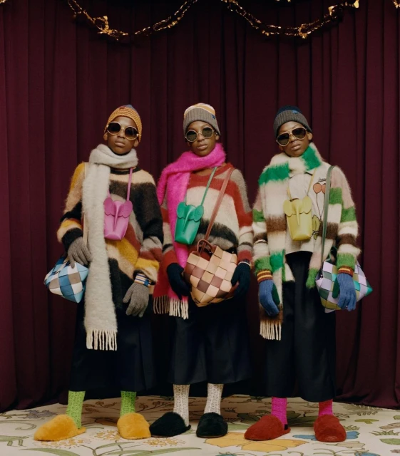 Three young, androgynous people in striped sweaters, hats, and sunglasses pose indoors with purses. Colorful accessories and a curtained backdrop.