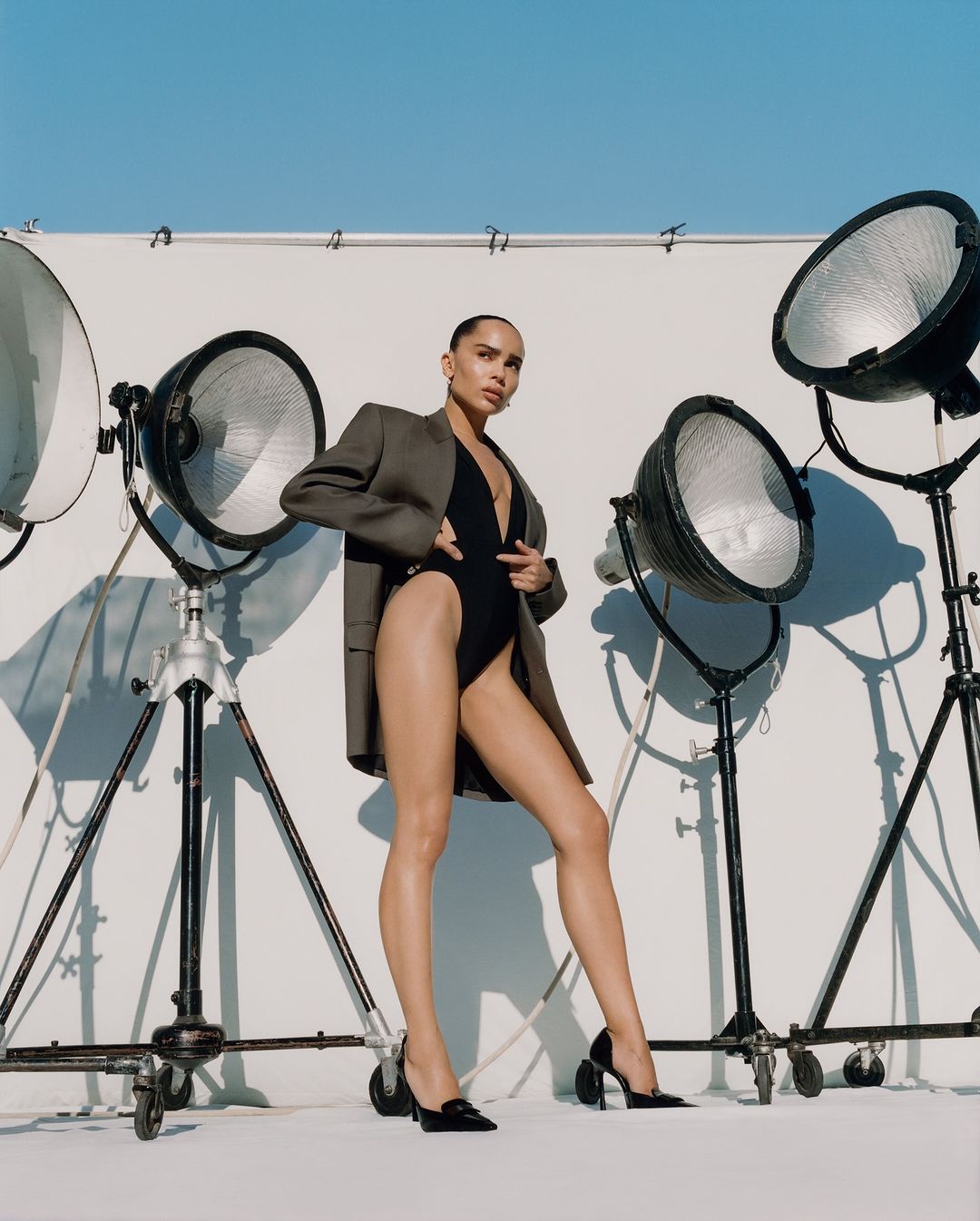 Attractive adult woman in a black bodysuit and blazer poses in front of lighting equipment on a white background in a studio setting.