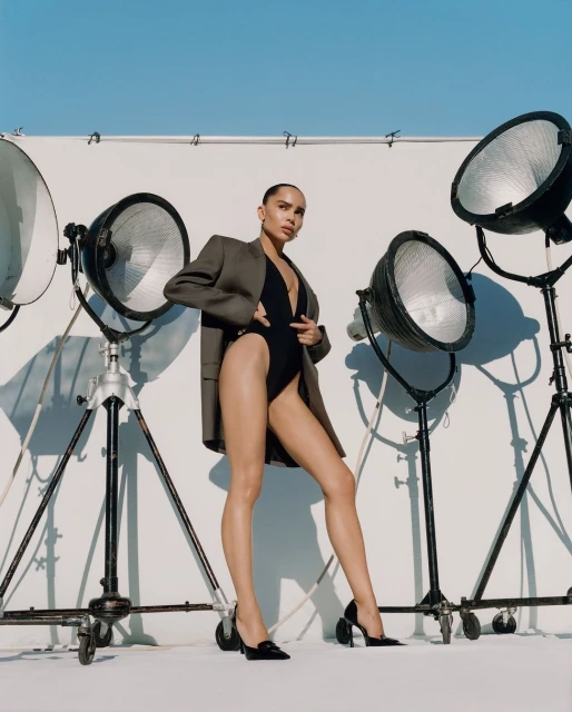 Attractive adult woman in a black bodysuit and blazer poses in front of lighting equipment on a white background in a studio setting.