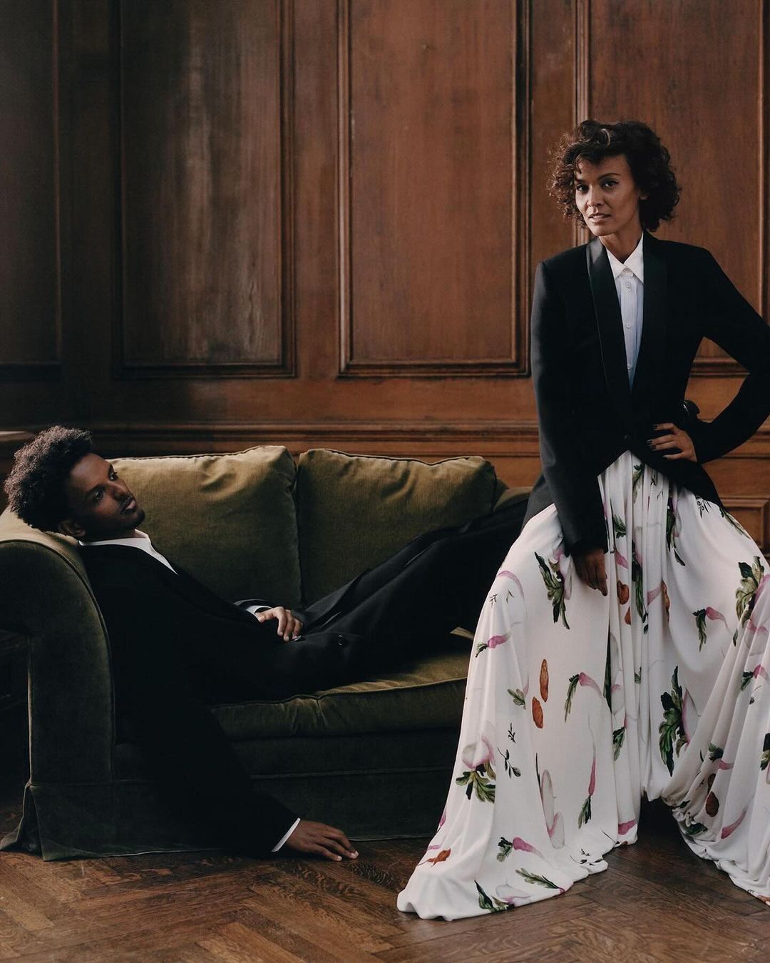 A woman standing in a patterned white skirt and black blazer next to a man reclining on a green velvet sofa.