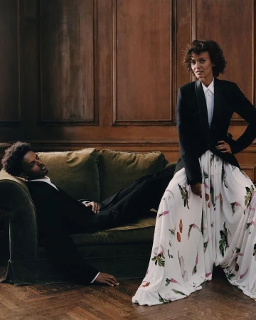 A woman standing in a patterned white skirt and black blazer next to a man reclining on a green velvet sofa.