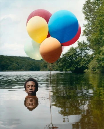 Portrait of a black male in a lake with closed eyes holding a colorful balloon bunch, trees and sky in background. Conceptual outdoor scene.