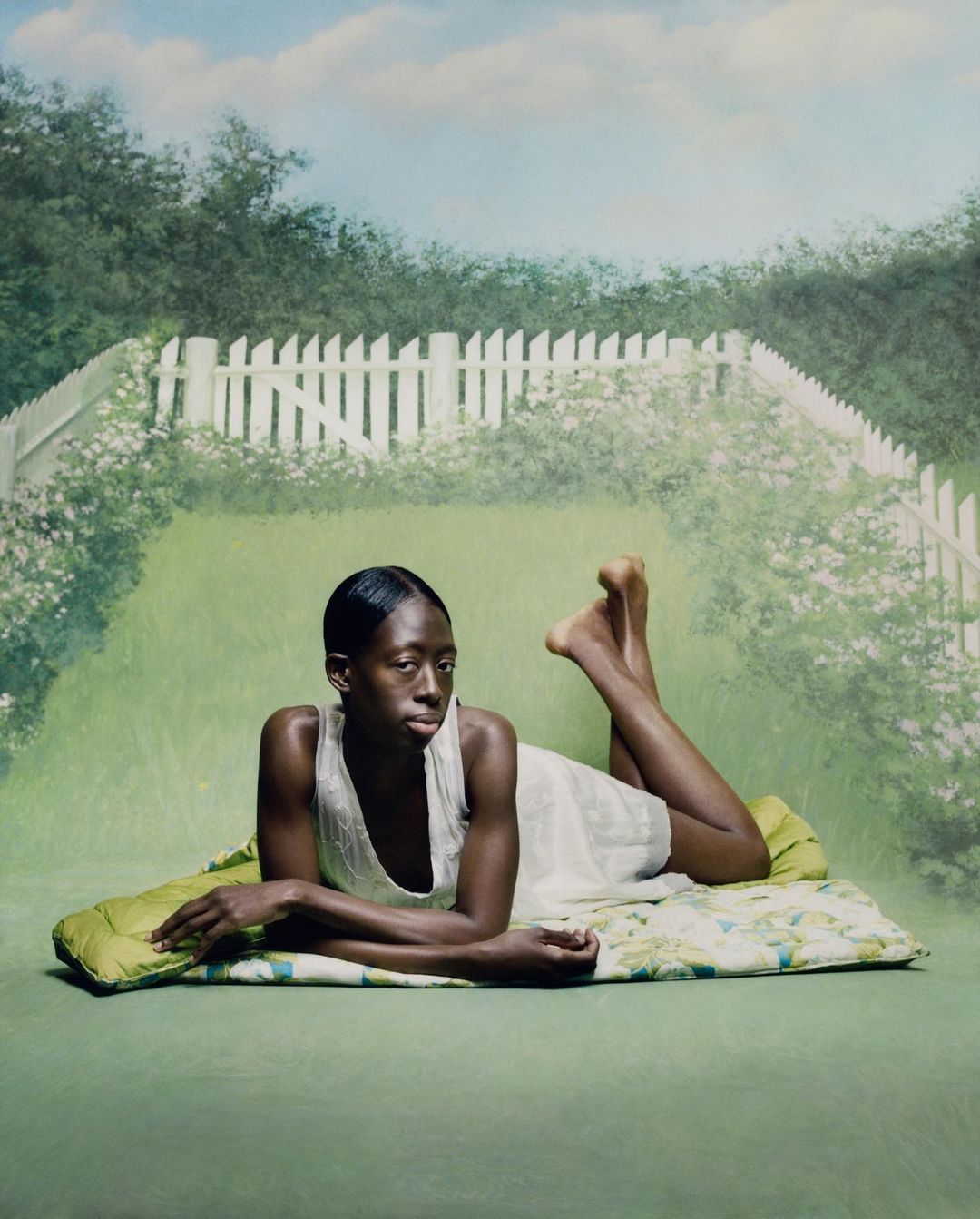 Woman lounges on a patterned pad against a hand-painted backdrop of a green lawn, white picket fence, and blue sky with light clouds.
