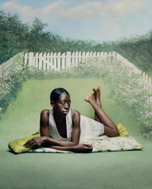 Woman lounges on a patterned pad against a hand-painted backdrop of a green lawn, white picket fence, and blue sky with light clouds.