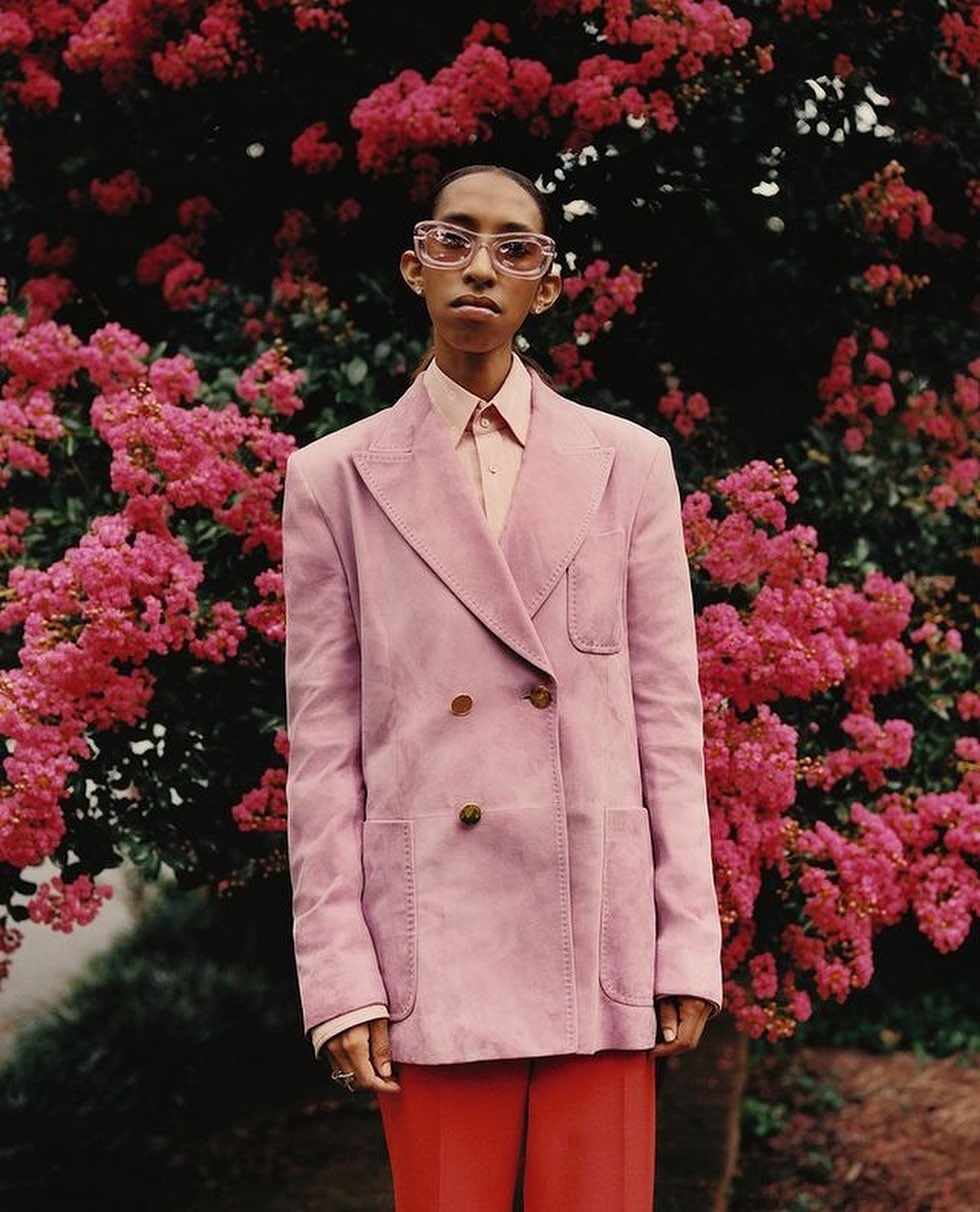 A person poses in a pink double-breasted blazer and red pants against a backdrop of bright pink flowers, presenting a fashion-forward look.