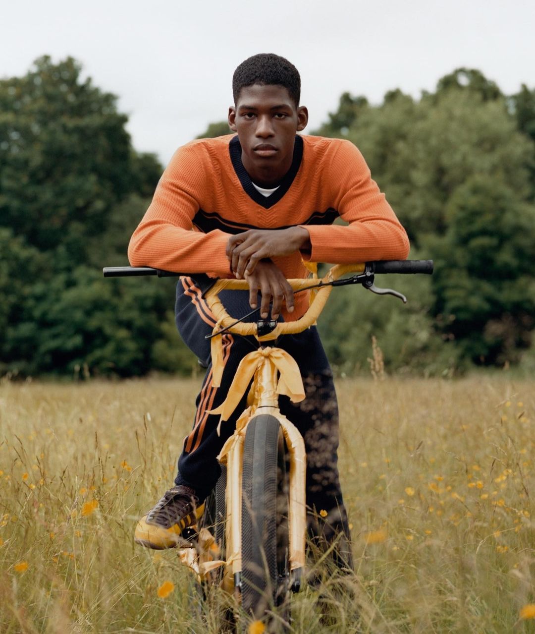 A dark-skinned man poses with a yellow bicycle in a grassy field with trees in the background, wearing an orange sweater.