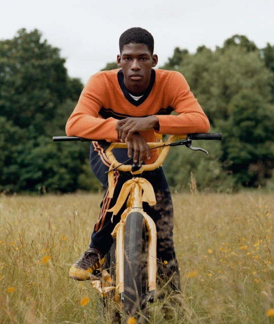 A dark-skinned man poses with a yellow bicycle in a grassy field with trees in the background, wearing an orange sweater.