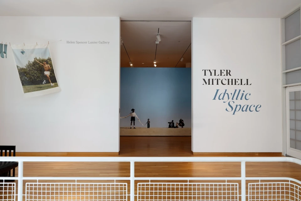 A minimalist art gallery view featuring artwork, exhibition titles, and white walls, complemented by a light wood floor and natural light.