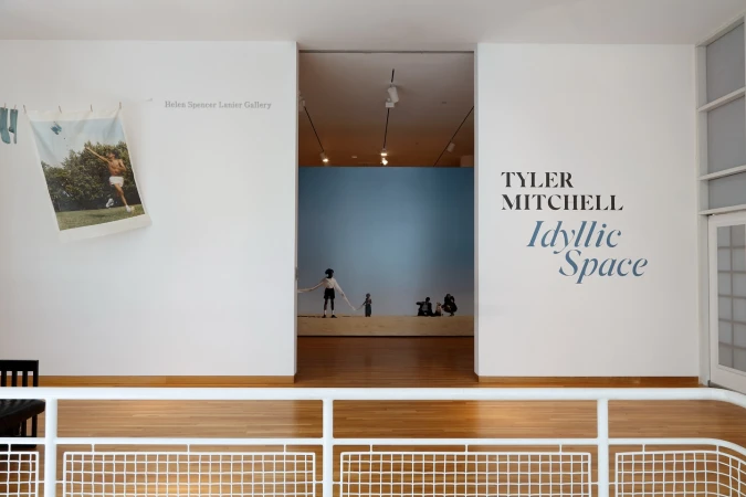 A minimalist art gallery view featuring artwork, exhibition titles, and white walls, complemented by a light wood floor and natural light.