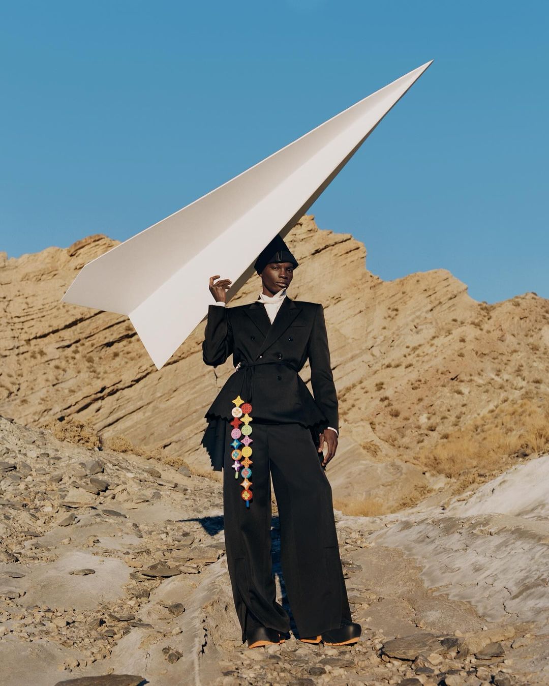 Fashionable woman in a black suit holds a large paper airplane outdoors, standing against a backdrop of a clear sky and rock formations.