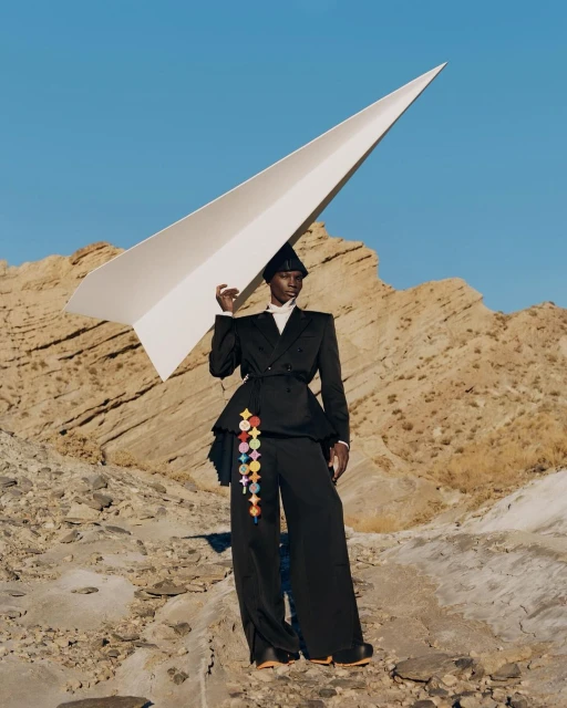 Fashionable woman in a black suit holds a large paper airplane outdoors, standing against a backdrop of a clear sky and rock formations.