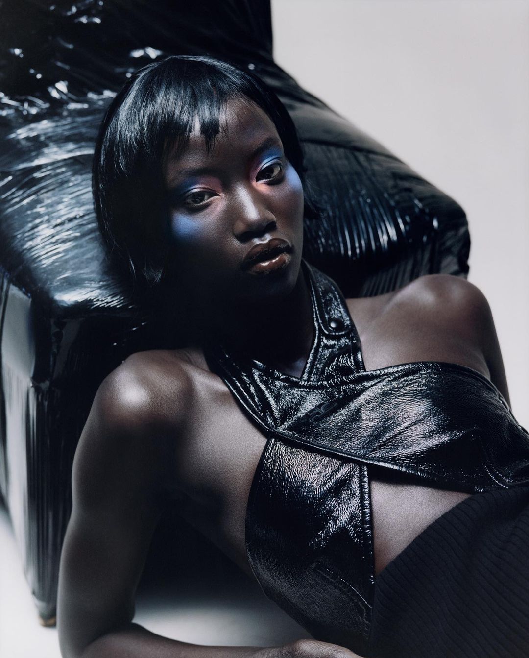 Portrait of a woman with blue and red eyeshadow and dark hair, laying on a black shiny leather chair in a studio, beauty shot.