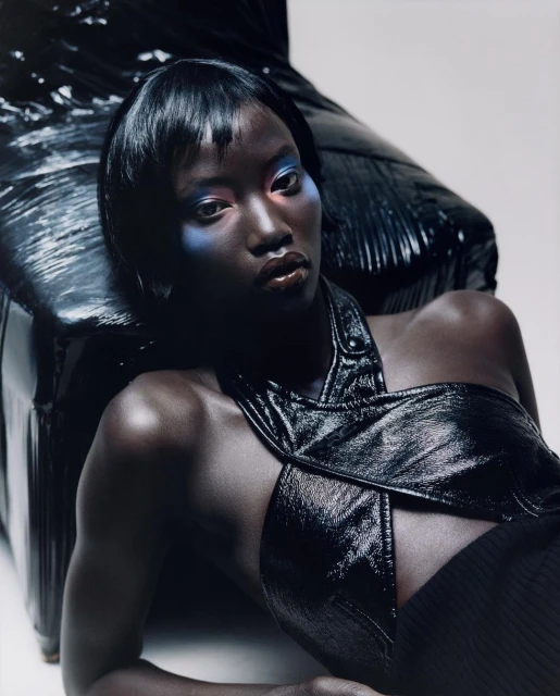 Portrait of a woman with blue and red eyeshadow and dark hair, laying on a black shiny leather chair in a studio, beauty shot.