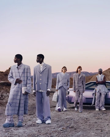 Five fashion models in light purple suits pose outdoors on a desert landscape near a matching purple car in a fashion editorial shoot.