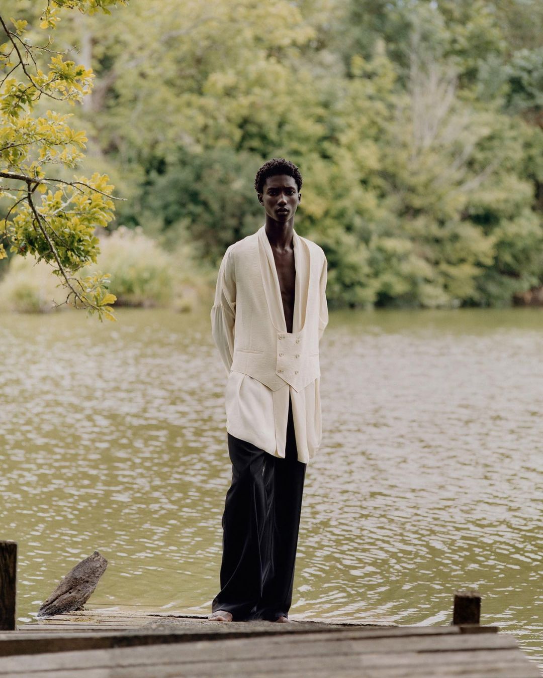 A slender African-American man in a cream-colored waistcoat and black pants poses on a wooden dock with a lake and trees in the background.