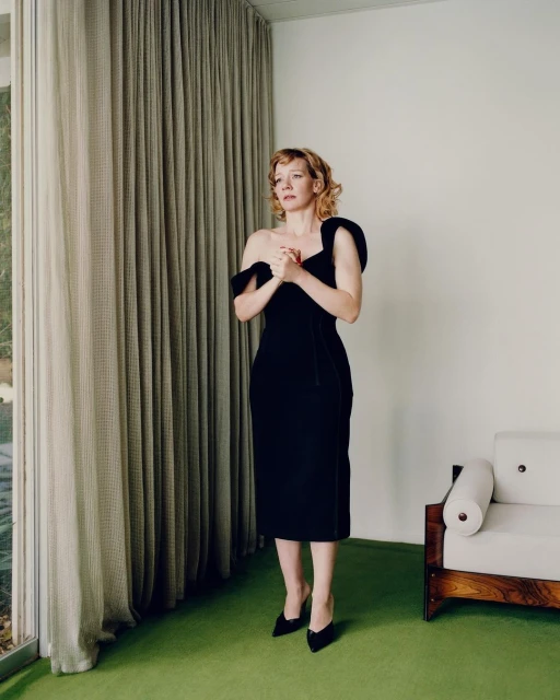 Woman in black dress stands in interior, posed for full-body portrait, natural light through window, neutral background, and minimalist decor.