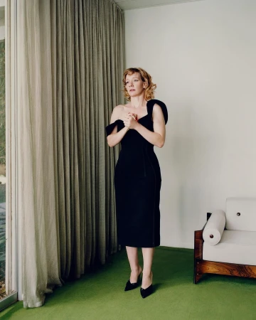 Woman in black dress stands in interior, posed for full-body portrait, natural light through window, neutral background, and minimalist decor.