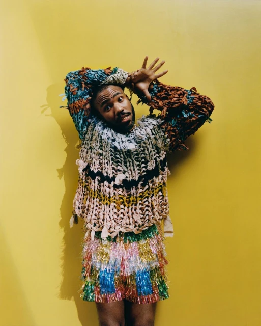 Adult male poses against a yellow background, wearing a hand-crafted colorful sweater and fringed skirt, with hands raised above his head.