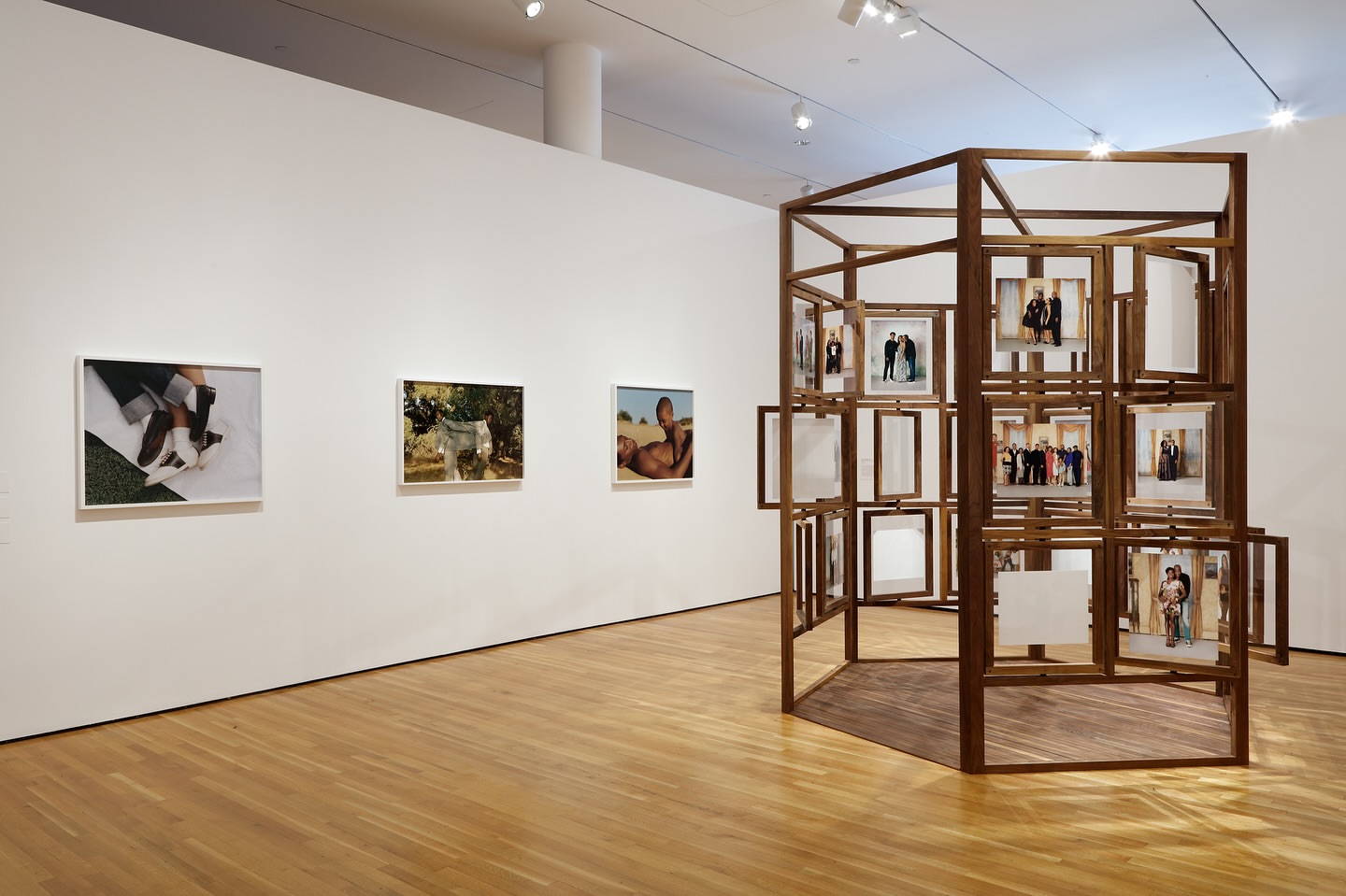 Art gallery interior with framed photos on a white wall and a wooden geometric photo display structure on a wood floor.