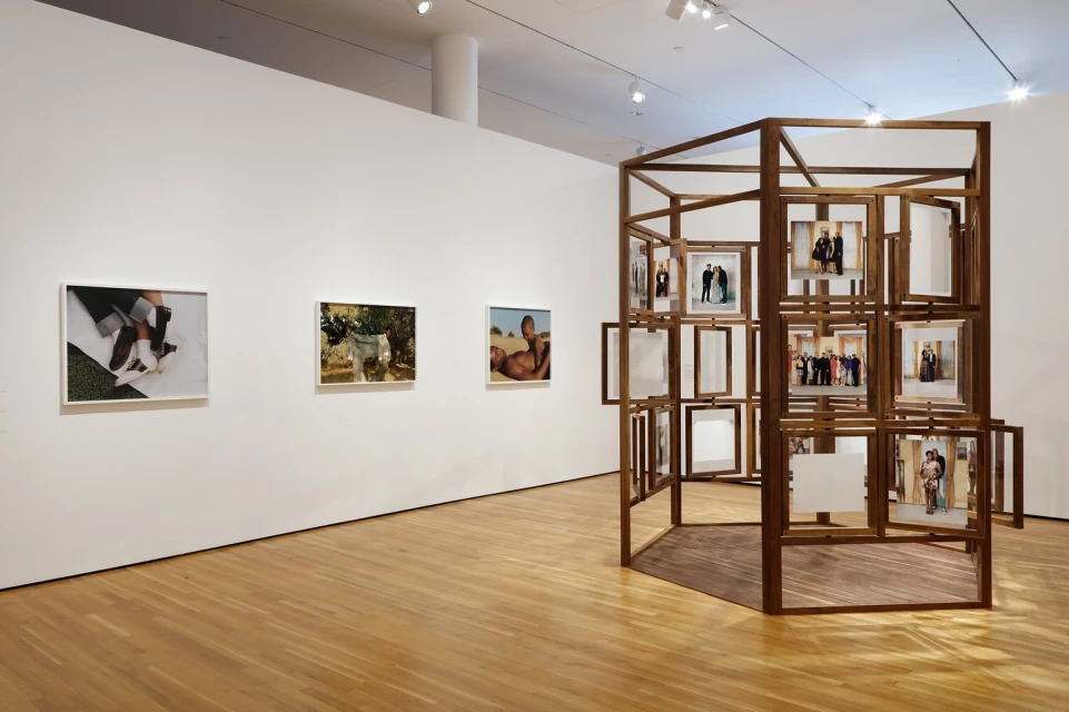 Art gallery interior with framed photos on a white wall and a wooden geometric photo display structure on a wood floor.