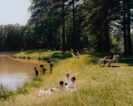 Group of Black people enjoying a sunny day by a lake in a grassy meadow surrounded by tall trees, photographed by Tyler Mitchell.