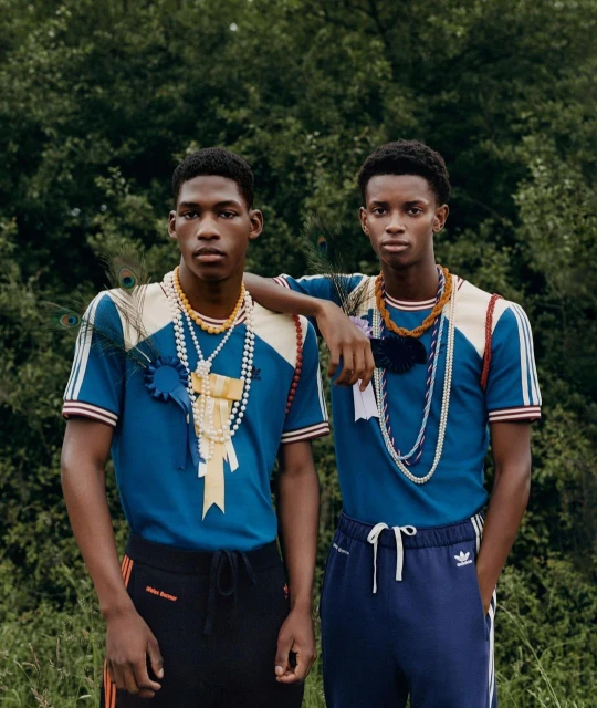 Two stylish Black men pose outdoors wearing matching blue t-shirts and dark pants with decorative accessories, exuding a fashion-forward vibe.