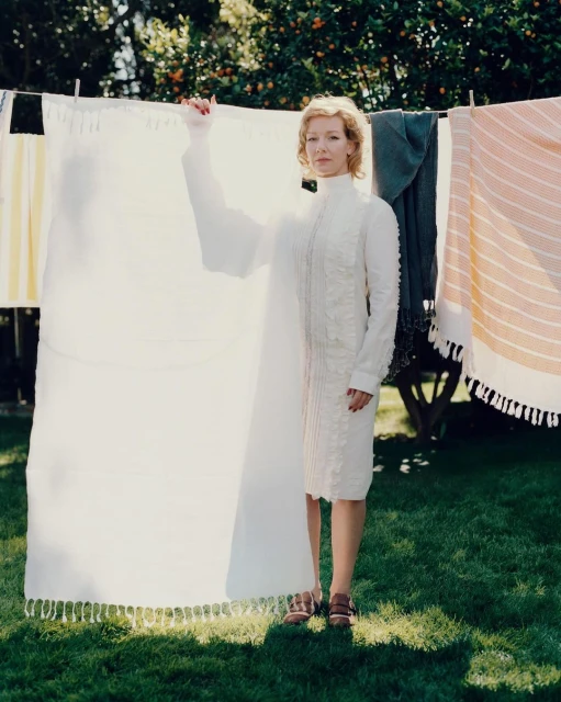 Woman in white dress standing near a clothesline with white and striped towels hanging on a sunny day in a grassy backyard.
