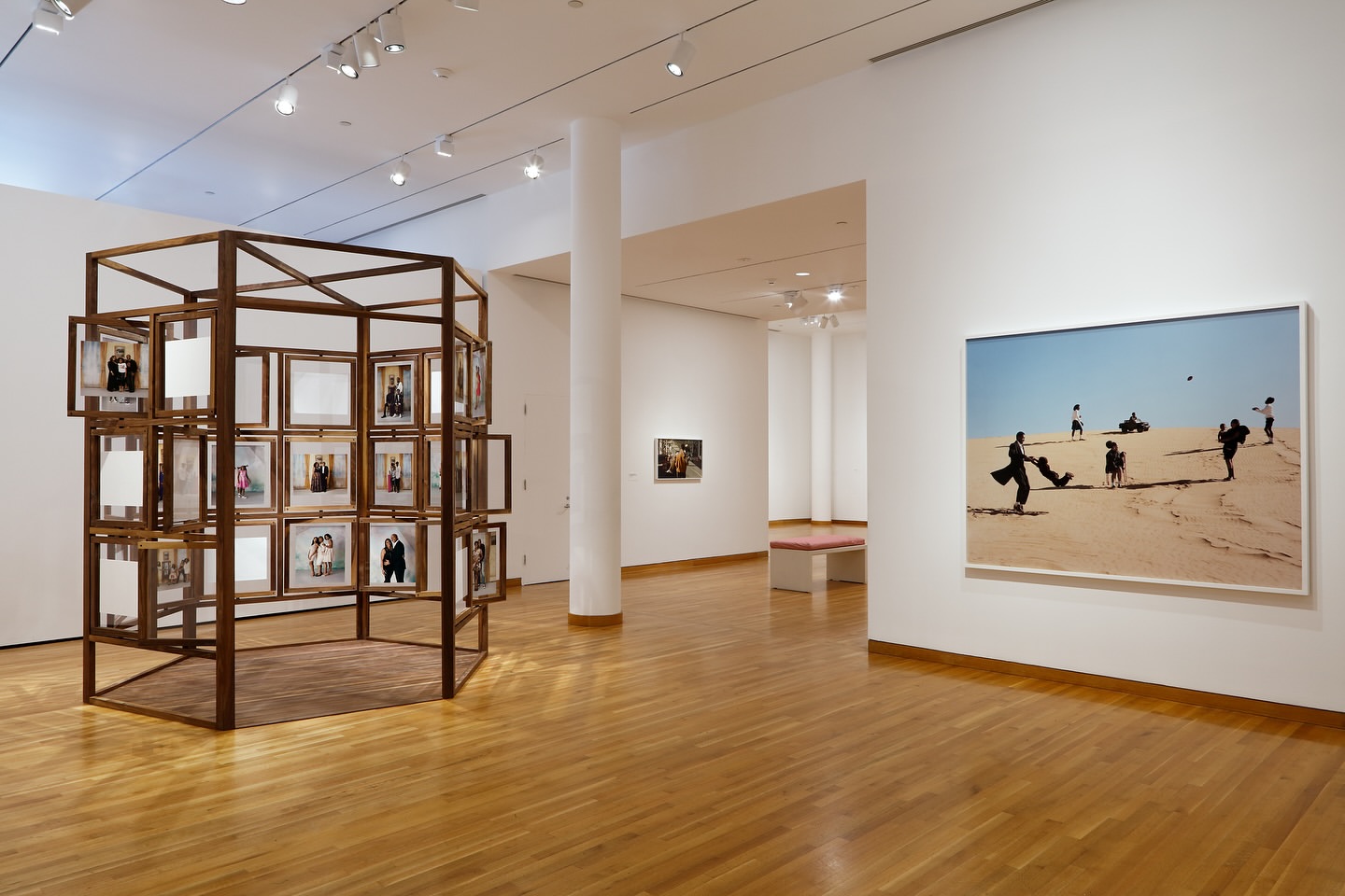 Art gallery interior featuring a framed photograph of figures on sand and a wooden installation with framed portraits.