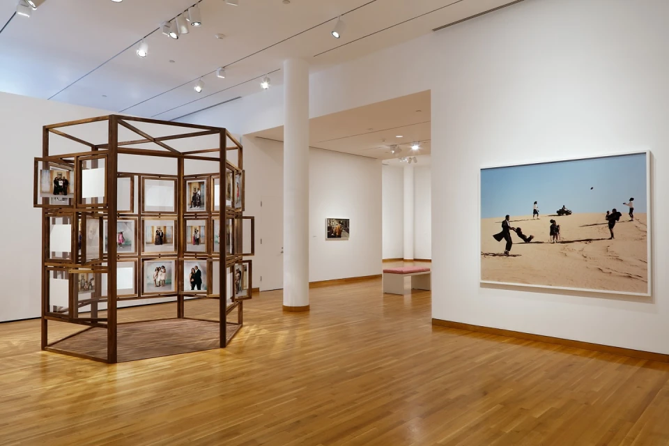 Art gallery interior featuring a framed photograph of figures on sand and a wooden installation with framed portraits.