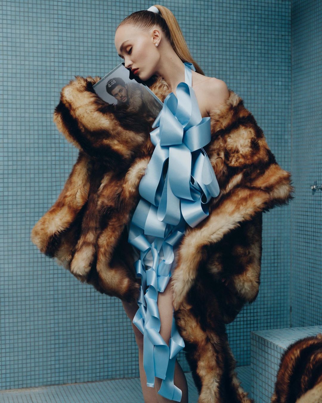 Model wearing a brown fur coat and blue ribbon halter top holding a magazine in a tiled bathroom setting