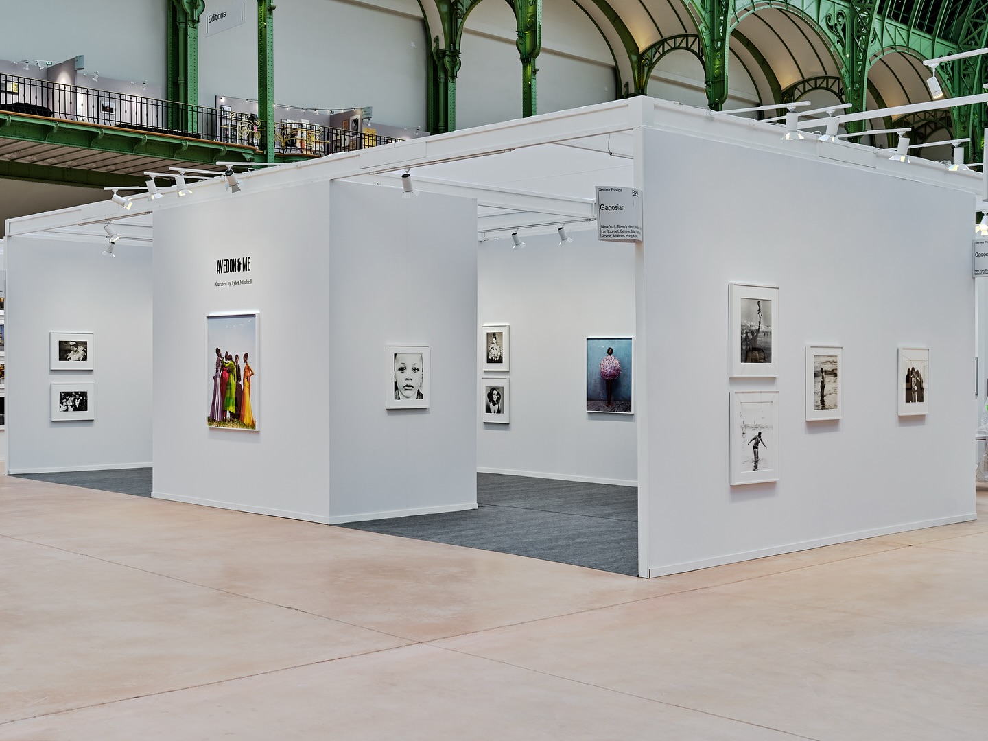 Interior view of art exhibition featuring photographs and white display booths beneath industrial architecture with natural lighting.