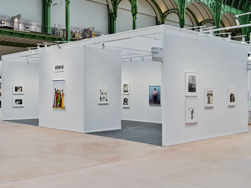 Interior view of art exhibition featuring photographs and white display booths beneath industrial architecture with natural lighting.