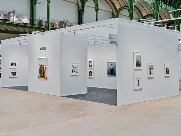 Interior view of art exhibition featuring photographs and white display booths beneath industrial architecture with natural lighting.