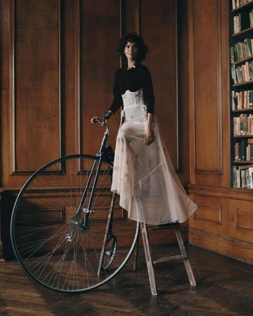 Adult woman poses next to a vintage bicycle in an elegant room with a library. She wears a long skirt and a corset.