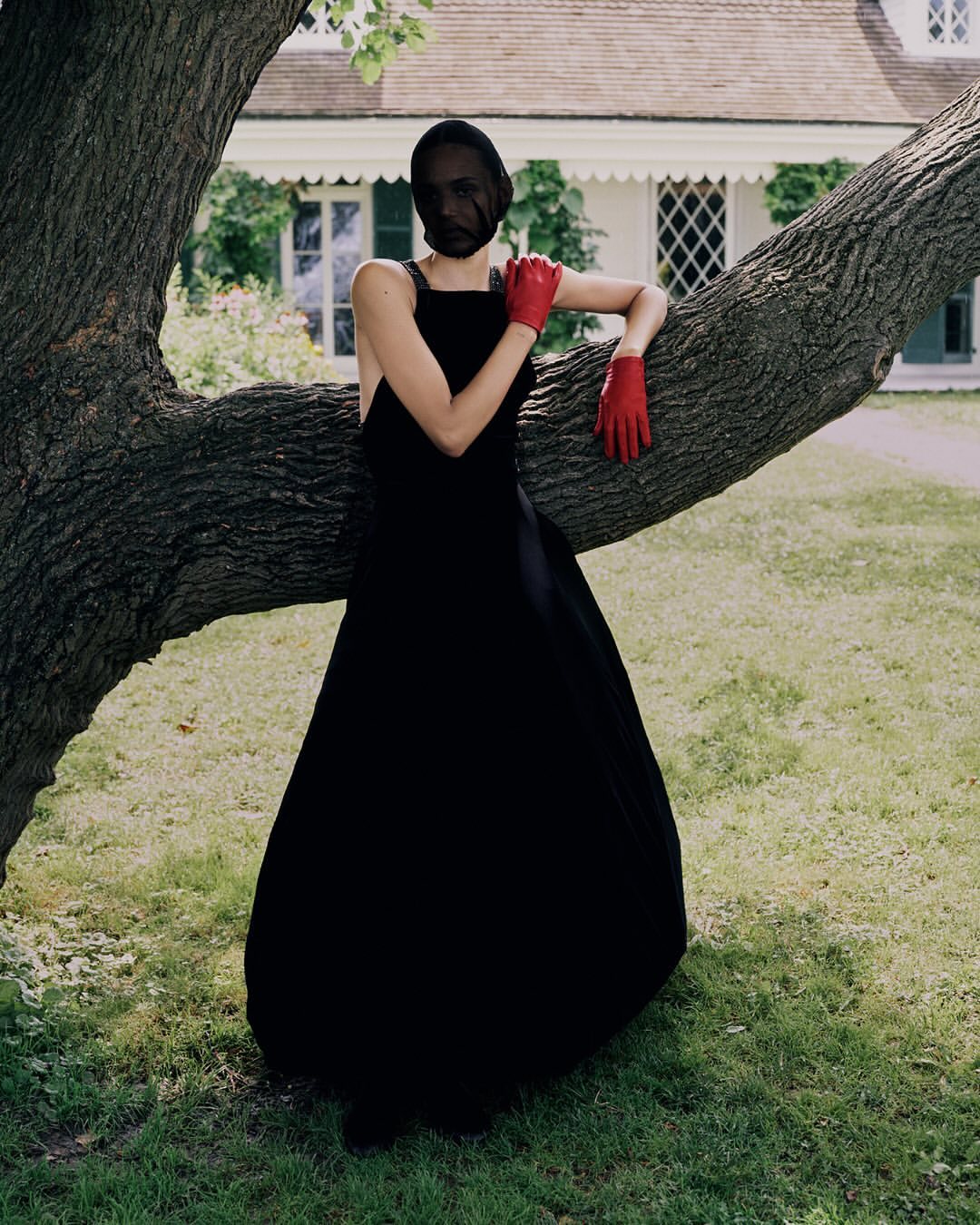 A woman in a black dress and red gloves leans against a tree in a grassy yard with a white house in the background on a sunny day.