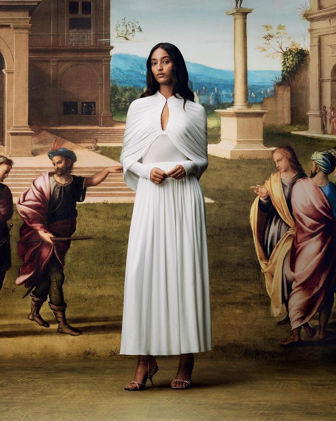 Elegant woman in long white dress poses in front of a painted backdrop with classical architecture and figures, under soft light.